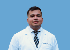 Studio headshot of a male dentist wearing a white lab coat and tie against a blue background